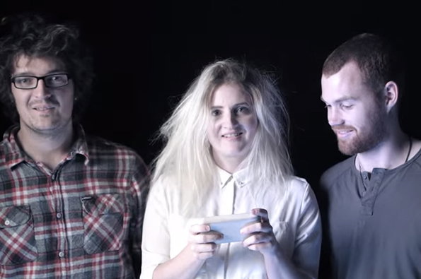 Three people stand against a black background. The person in the centre holds a smartphone and looks at the camera, while the other two look on.