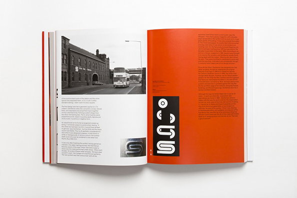 An open book shows a spread with a photograph of a brick building and a white bus on a city street. The opposite page is bright orange and features a black vertical strip containing a white, stylised “S” logo.