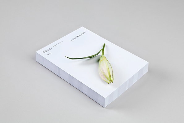 A white notepad rests on a plain grey surface with a single white lily bud laid across the top page. The header reads “Critical Mass Studio” with contact details printed in a smaller font.