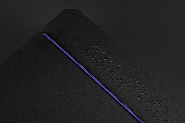 A close-up of a matte black book or folder with a thin purple elastic band. Braille dots are embossed into the cover.
