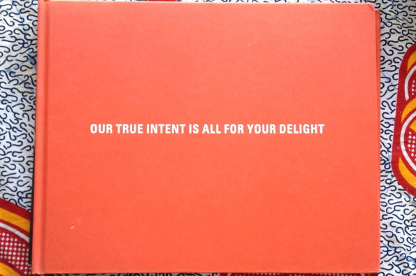 A bright orange book cover with the words “OUR TRUE INTENT IS ALL FOR YOUR DELIGHT” printed in white across the centre. The book rests on a patterned fabric with red and blue details.