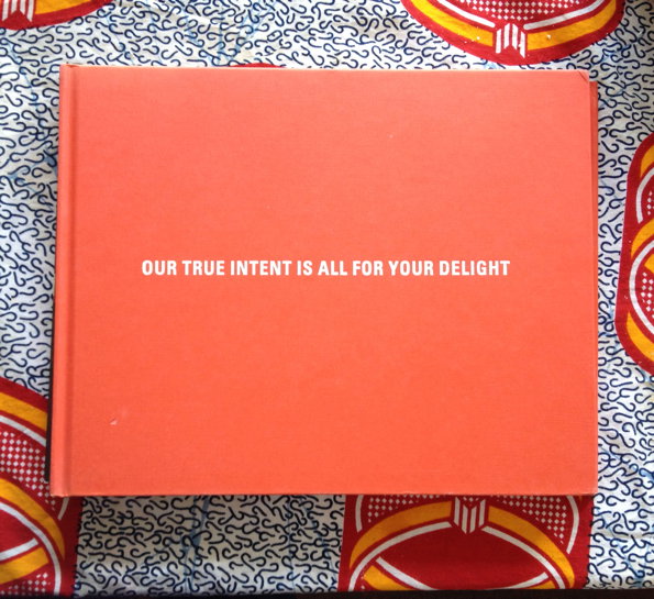 A bright orange book lies flat on a patterned fabric of blue, red and yellow. Centred on the cover, white text reads “OUR TRUE INTENT IS ALL FOR YOUR DELIGHT”.
