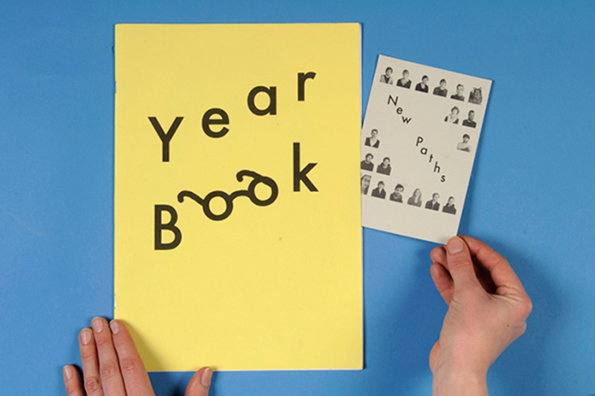 A yellow book cover reads “Year Book” in black typography, with the letters in “Book” styled to look like a pair of glasses. A person’s hands hold the book and a smaller white card that reads “New Paths” alongside small portrait photos.