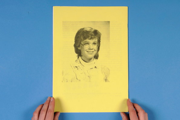 Two hands hold a bright yellow sheet of paper against a blue background. The page features a monochrome portrait of a smiling person wearing glasses.