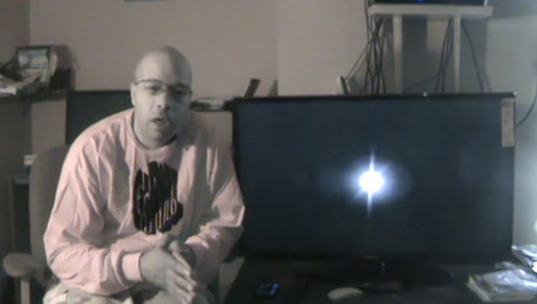 A person wearing glasses and a pink sweatshirt sits in a room next to a large computer monitor. They are gesturing with their hands while looking toward the camera.