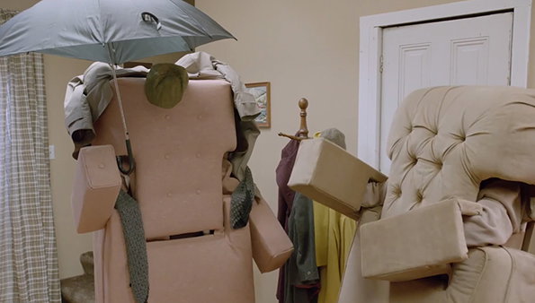 Two upholstered armchairs are dressed in clothes to resemble people. One chair wears a beige jacket and holds a grey umbrella, while the other wears a yellow garment.