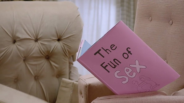 A pink book titled “The Fun of Sex” rests on the arm of a beige tufted armchair. Another book is partially visible behind it.