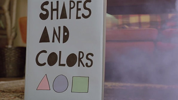 A close-up of a book cover with the title “SHAPES AND COLORS” in bold, hand-drawn black lettering. Below the text are three simple shapes: a pink triangle, a purple circle and a green square.