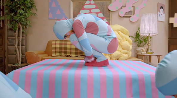 A person in a blue and pink patterned bodysuit crouches on a pink and blue striped platform. They are surrounded by a surreal room with pastel shapes on the walls and a yellow textured sculpture.