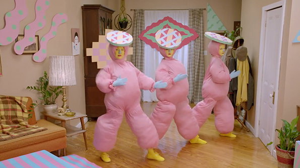 Three people wear oversized, puffy pink costumes and wide, patterned hats. They stand in a living room with a brown sofa and a white pleated lamp.