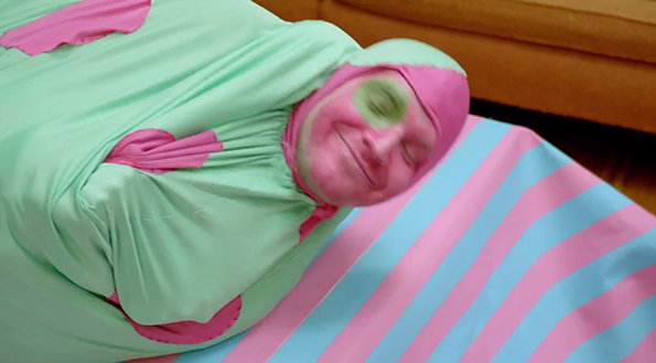 A person wrapped in a tight, mint-green fabric costume lies on a pink and blue striped surface. Their face is covered in pink and green makeup, and they are smiling with their eyes closed.