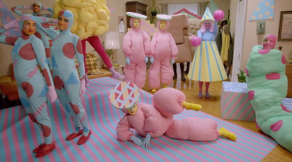 A group of people wear oversized, colourful costumes in a brightly lit room. The outfits include padded pink suits, blue patterned bodysuits, and a large green caterpillar shape.