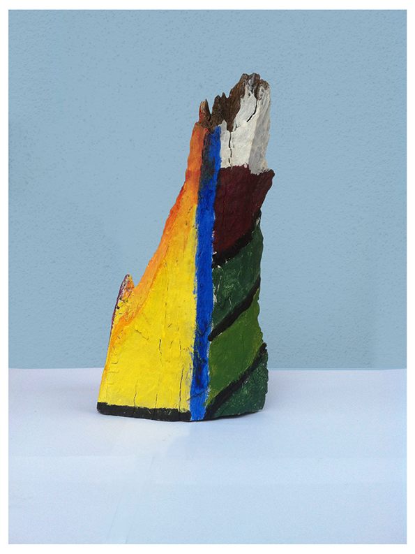 A sculpture made from a jagged piece of weathered wood, painted in bold vertical stripes of yellow, blue, green and red. The top of the piece remains unpainted, showing the natural grey grain of the wood against a pale blue background.