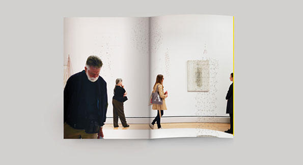 An open book shows a photograph of people visiting an art gallery. Several figures stand in a white room, looking at a framed piece of art on the wall.