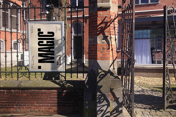 A white sign hangs from a black wrought iron fence in front of a red brick building. The sign features bold, black typographic text that reads “MAGIC”.
