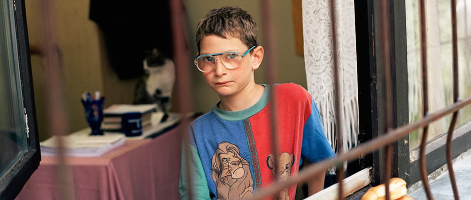 A child wearing large glasses and a colourful T-shirt looks directly at the camera. They stand behind a metal railing, with a room containing a desk and a cat visible behind them.