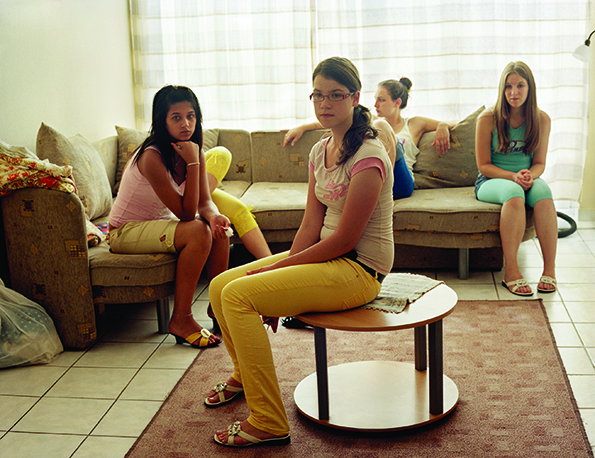 Four people sit in a living room, looking towards the camera with neutral expressions. One person sits on a small round table in the foreground, while the others are positioned on a beige sofa.