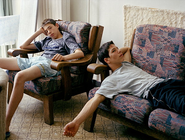 Two people lounge in patterned armchairs within a sparsely furnished room. One person sits upright with their head resting on their hand, while the other reclines across a second chair.