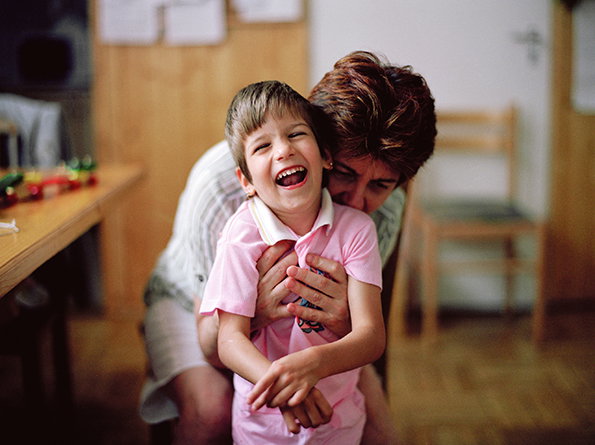 A person hugs a laughing child from behind. The child wears a pink polo shirt and leans back with their mouth open in a wide smile.