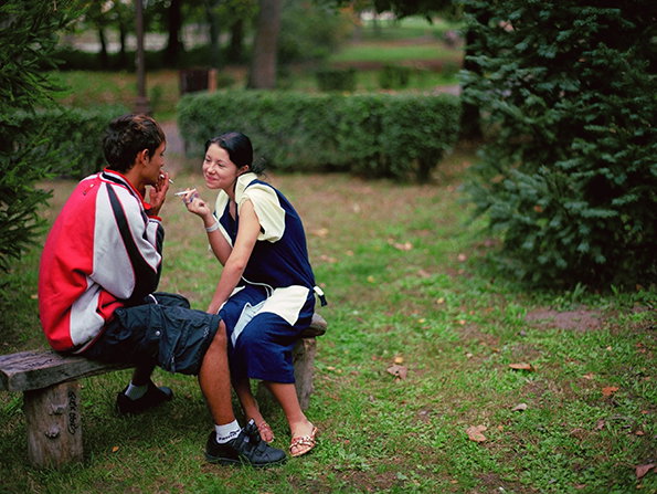 Two people sit on a wooden bench in a green park, facing each other and talking. One person wears a red and white jacket, while the other wears a blue and yellow outfit.