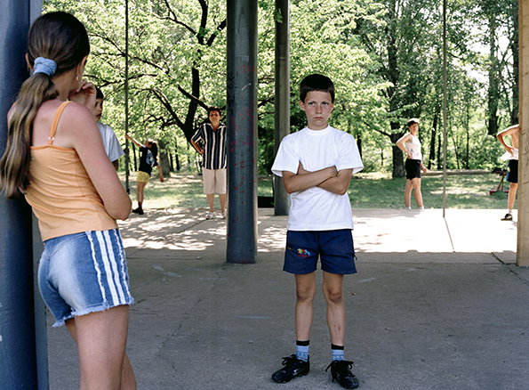 A child in a white T-shirt and blue shorts stands with arms crossed, looking directly at the camera. Other children stand in the background under a canopy of green trees.