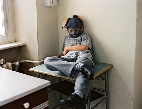 A child sits on a blue table in a plain room, wearing a grey T-shirt and trousers. Their face is covered in dark grey makeup and they wear a matching grey and orange headpiece.
