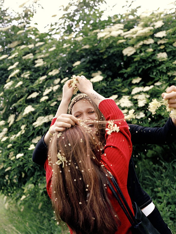 Two people laugh and embrace in front of a dense hedge of white flowers. One person wears a red sweater and has small white blossoms scattered through their long brown hair.