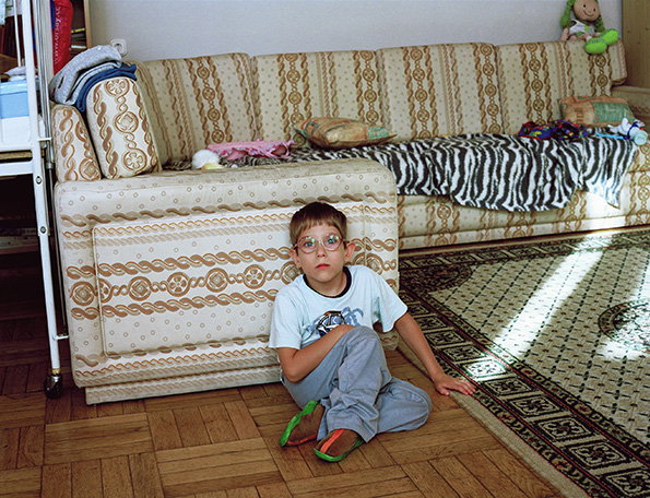 A child wearing glasses and a light blue T-shirt sits on a wooden floor, leaning against a patterned sofa. A zebra-print throw and various cushions are scattered across the seating.