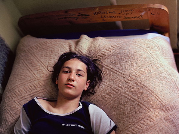 A person lies on a bed covered in a textured beige blanket, looking directly at the camera. The wooden headboard behind them is scrawled with handwritten messages, including “I love you”.