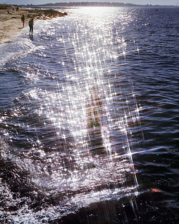 Bright sunlight glints off the surface of dark blue water, creating a shimmering path of white light. Two people stand on the sandy shoreline in the distance.