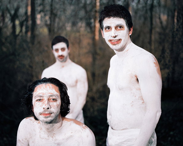Three people stand in a wooded area, their bodies and faces covered in thick white paint. They look towards the camera with neutral expressions.