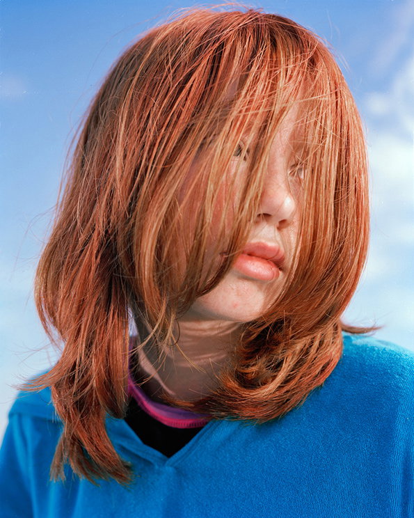 A person with long, reddish-orange hair that partially covers their face. They wear a bright blue top against a clear blue sky.