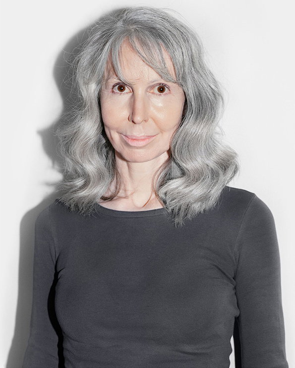 A person with wavy grey hair looks directly at the camera. They wear a plain dark grey long-sleeved top against a white background.