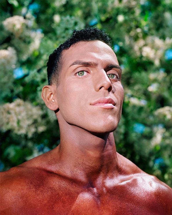 A person with short dark hair and light eyes looks slightly upwards with a neutral expression. They are shirtless, posed against a blurred background of green leaves and white blossoms.