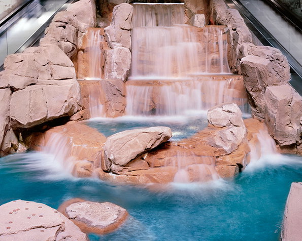 A tiered artificial waterfall flows over reddish-brown rocks into a bright blue pool. The water is captured with a soft, blurred effect as it cascades down the stone steps.