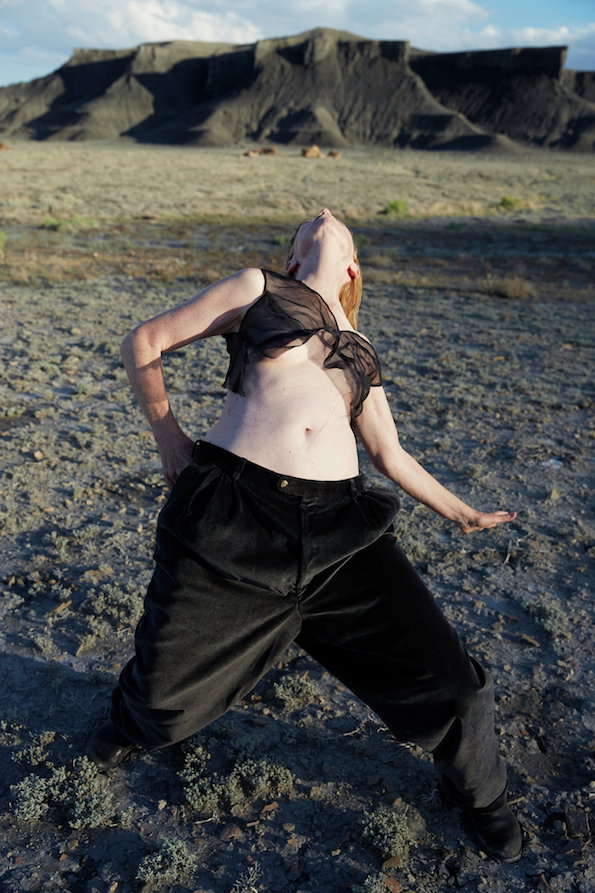 A person stands in a wide, arid landscape, leaning back with their head tilted towards the sky. They wear a sheer black top and oversized black trousers.
