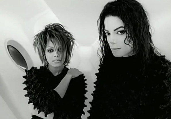 A black and white photograph of Janet and Michael Jackson. They both wear textured black clothing and pose against a minimalist, white architectural background.