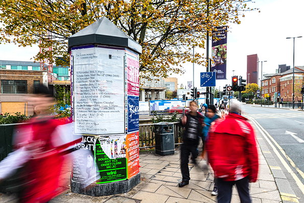 A street scene featuring a pillar covered in layered posters and flyers. Blurred figures of people walk past the pillar on a paved pavement under a tree with autumn leaves.