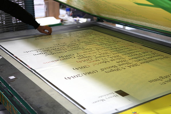 A person’s hand adjusts a large print inside a printing press. The print features a list of dates and names, including “Simone Forti, ‘Slant Board’ (1961/2014)”.