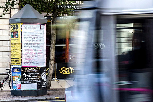 A grey cylindrical advertising pillar is covered in layered posters and flyers on a city street. A blurred bus passes in the foreground, partially obscuring a cafe in the background.