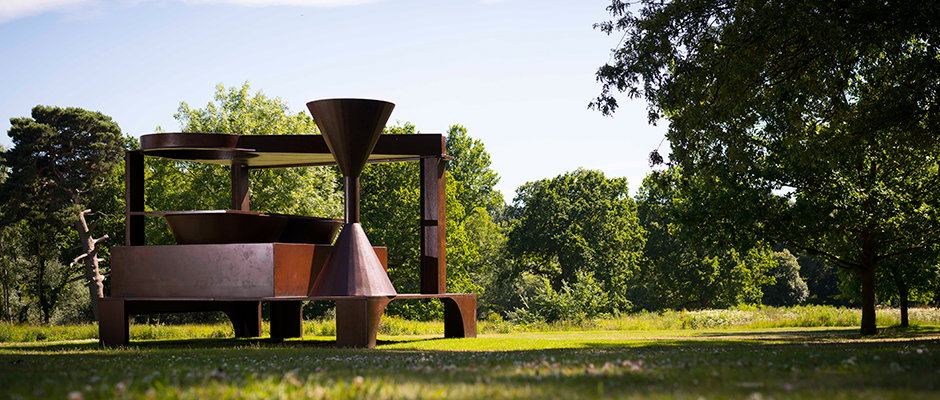 A large, rusted steel sculpture stands in a grassy field surrounded by green trees. The abstract work consists of heavy geometric plates and conical shapes arranged in a tiered structure.