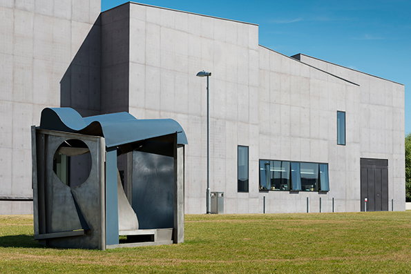 A concrete sculpture with a wavy blue roof sits on a green lawn. Behind it stands a large, minimalist concrete building with narrow windows under a clear blue sky.