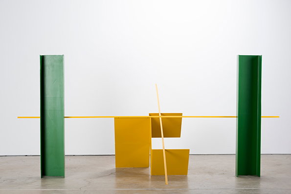 A sculpture consisting of green and yellow industrial steel beams against a white wall. Two green vertical beams flank a central arrangement of yellow beams, including a long horizontal bar and a vertical piece.