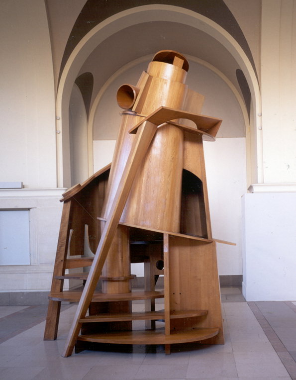 A large wooden sculpture of an abstract, tower-like structure stands in a gallery with arched ceilings. The piece features a mix of cylindrical and angular forms, including a set of stairs and several open apertures.