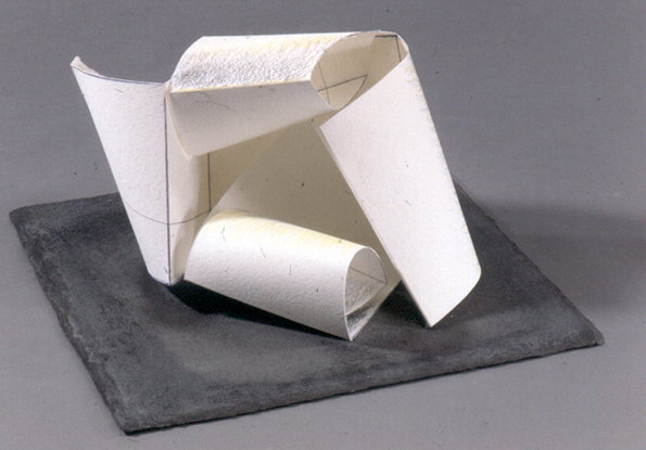 A white paper sculpture consisting of several folded and curled sheets. The abstract form sits on a dark grey rectangular base against a plain grey background.