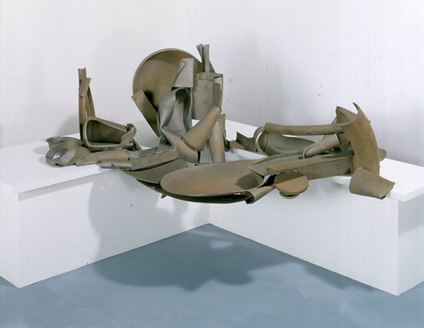 A large, abstract sculpture made of curved and folded beige sheets of metal. The pieces are arranged in a chaotic, overlapping pile across a white L-shaped plinth.