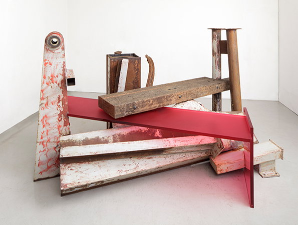 A sculpture made of weathered white and rusted steel beams arranged in a geometric pile. A flat, translucent red acrylic sheet cuts across the structure, resting on the beams.