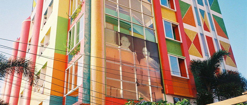 A brightly coloured building features a facade of bold yellow, red, green and blue panels. A large translucent image of two figures is displayed across several windows in the centre.