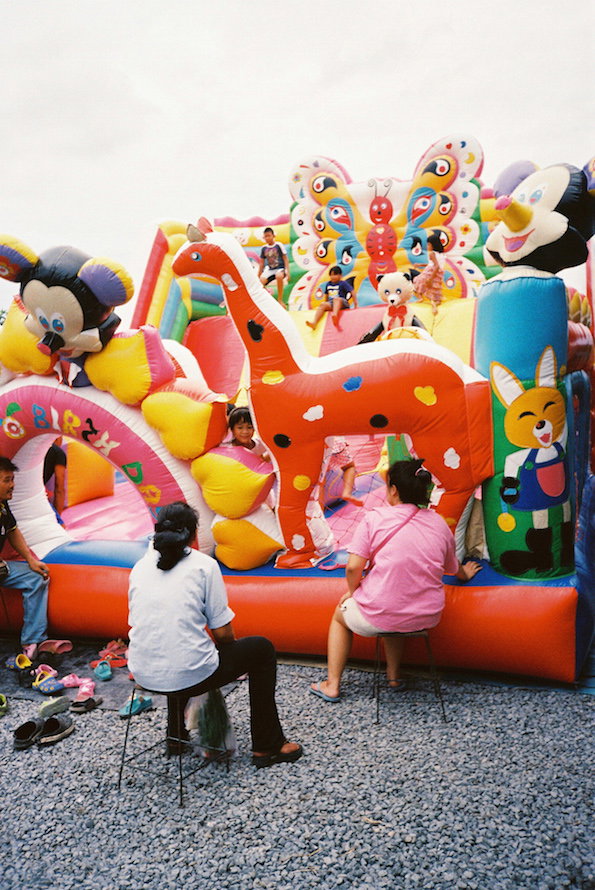 Children play on a large, colourful inflatable bouncy castle featuring a giant orange giraffe and a butterfly. Two people sit on folding chairs in the foreground, watching from a gravelled area.