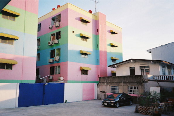 A multi-storey building is painted in wide horizontal stripes of pink, yellow, green and blue. A black car is parked in the concrete courtyard in front of the colourful facade.